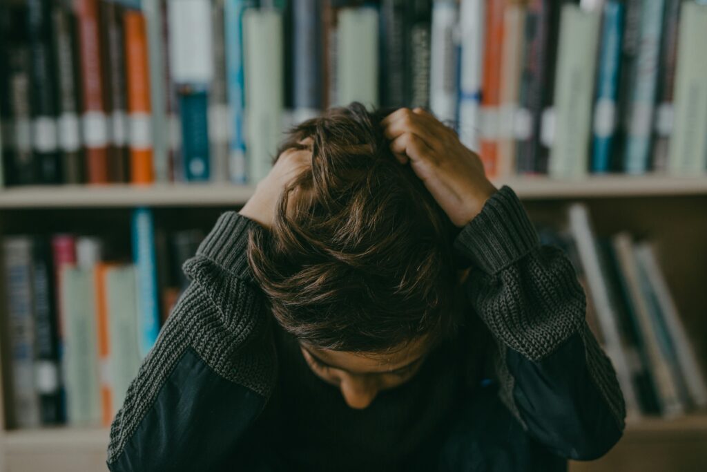 Here is a hospital-focused, SEO-optimized ALT text, written in a clinical yet human tone, perfect for the blog “Academic Stress in Students”: Alt Text: Student sitting in a library holding their head in stress, showing academic pressure, mental fatigue, and emotional overload commonly seen in students experiencing academic stress and anxiety.